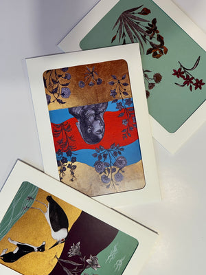 An assortment of original art greeting cards by Sumaira Tazeen, displaying various designs including a bird on a gold leaf background, floral patterns on a mint green surface, and a central portrait card, all reflecting a fusion of traditional and modern South Asian art styles.