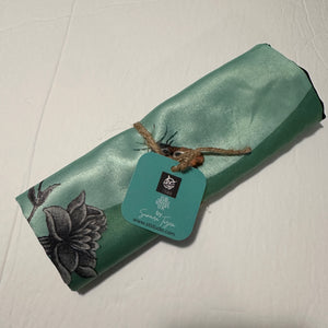 A rolled teal polyester miniature art cushion cover showing the soft sheen texture and Sumaira Tazeen artist label attached with jute twine.