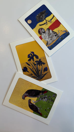 A set of three printed fine art greeting cards by Sumaira Tazeen arranged on a white surface; the top card depicts a figurative scene of a couple under a night sky, the middle card shows deep blue irises (flora), and the bottom card features two birds among green foliage (fauna), all rendered in a contemporary South Asian miniature painting style with gold accents.
