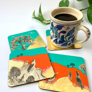 A blue and white floral coffee mug resting on a gold leaf coaster next to other hand-painted coasters showing bright orange and turquoise landscapes and bird motifs.