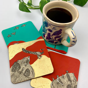 A lifestyle shot of a ceramic mug on a teal coaster, surrounded by scattered hand-painted coasters featuring birds in flight, a tall tree standing under a full moon, and birds perched on gilded hills.