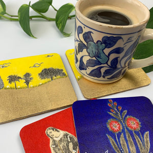 A white ceramic mug sits on a yellow landscape hand-painted coaster, with a second yellow coaster beside it and the blue and red ones layered nearby to show the complete set.