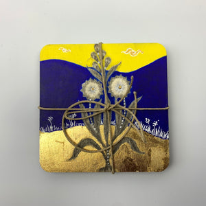 A close-up view of a hand-painted coaster presented in packaged form with a jute string tie featuring a flower with fine brushwork on a blue and yellow backdrop with gold leaf accents.