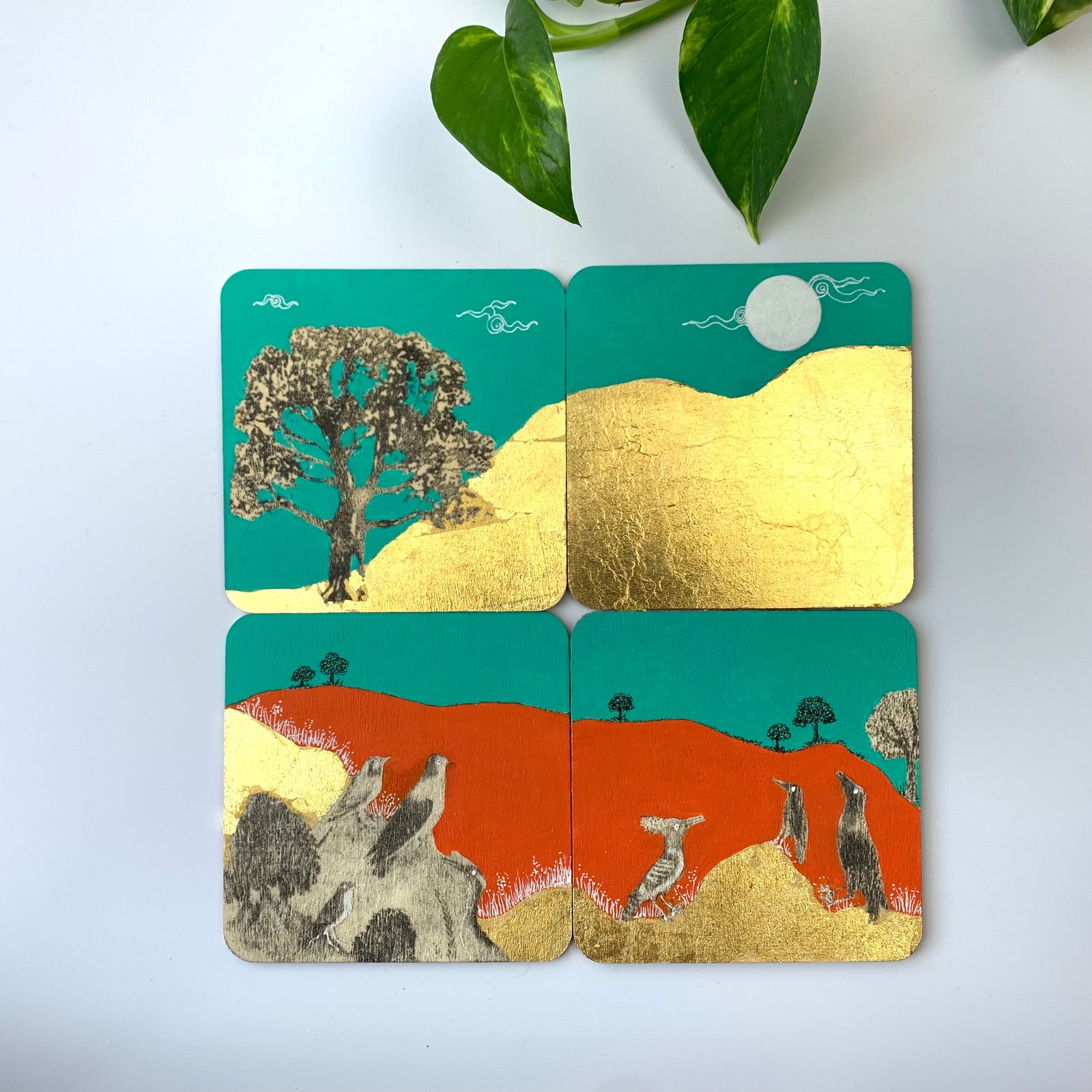 A set of 4 hand-painted coasters featuring the Bird's Choir theme with gold leaf hills, vibrant orange and turquoise backgrounds, and motifs of trees and birds.
