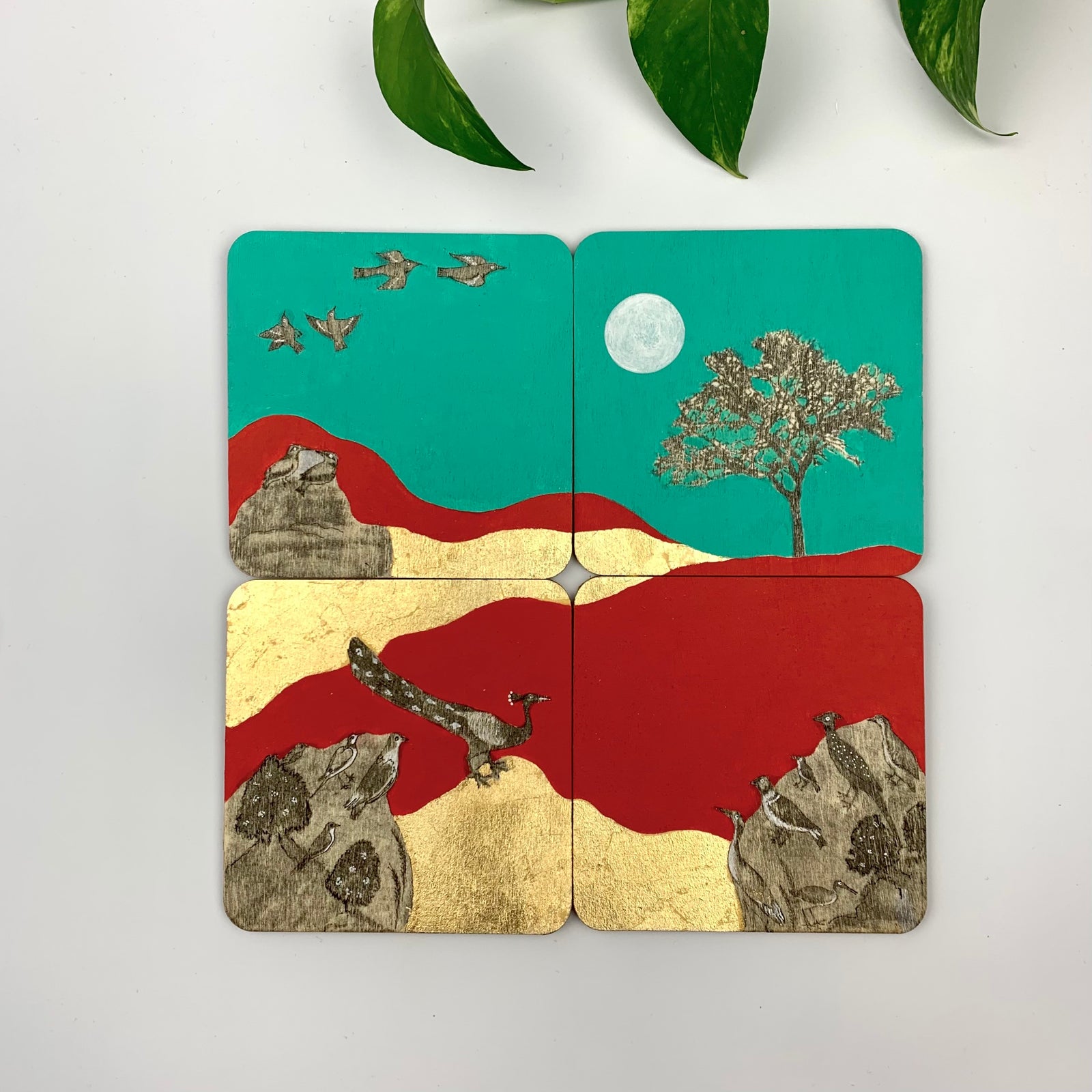 A set of four hand-painted coasters showing a continuous landscape of royal peacocks, flying birds, and gilded trees against teal and crimson backgrounds.