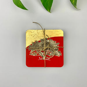 Top-down view of a packaged gift set of coasters featuring a tree on a red and gilded background, secured with a  twine bow for a handmade appeal.