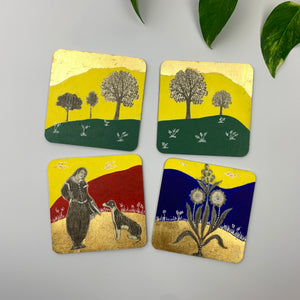 Four scattered hand-painted coasters featuring gold leaf hills and miniature art motifs, including a woman with a dog and vibrant floral botanicals.