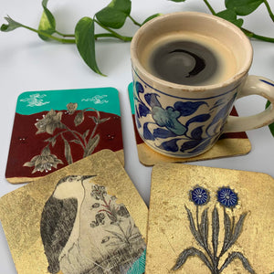 A lifestyle shot of a blue and white ceramic coffee mug resting on a hand-painted coaster with gold leaf detailing. The surrounding hand-painted coasters display intricate Mughal-style motifs including a bird and blooming flowers set against vibrant turquoise, deep red, and shimmering gold backgrounds.