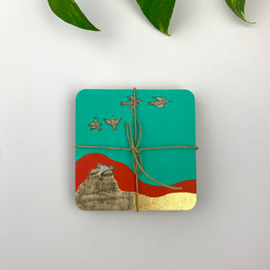 A top-down view of a single hand-painted coaster in gift-ready form, featuring birds flying over a crimson hill against a vibrant teal sky.