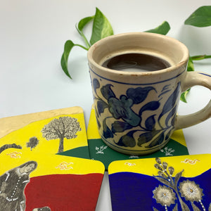 A lifestyle shot of a cofee mug resting on a forest green coaster, surrounded by scattered hand-painted coasters showing deep blue, crimson and yellow designs.